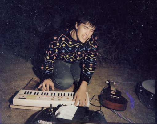 Very grainy polaroid of Jon kneeling on a rectangular piece of wood while playing a casio keyboard that is sitting on the ground and singing at night in a park with other musical instruments scattered around.