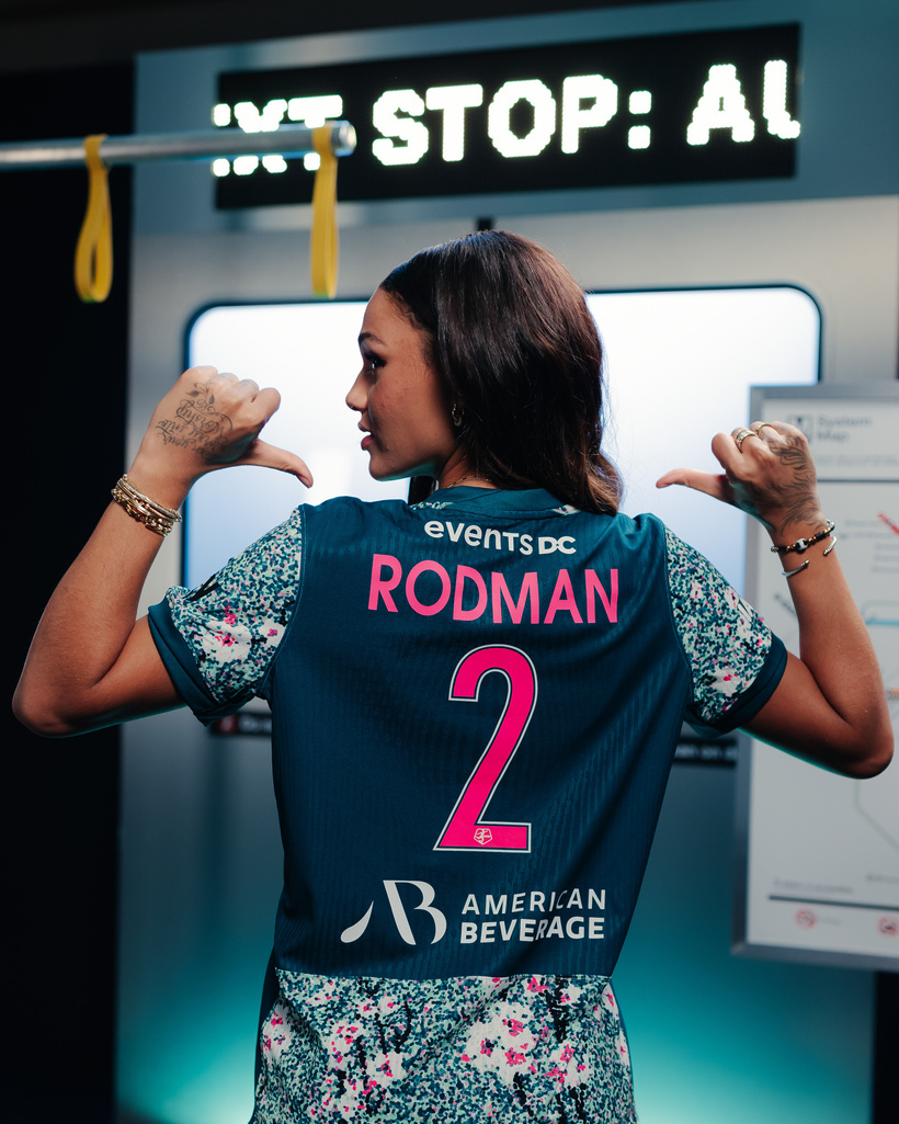 NWSL star Trinity Rodman modeling her subway series uniform for the Washington Spirit. It is Blue with pink and white abstract cherry blossom imagery. Her name RODMAN and number 2 appear in large pink lettering on a blue background.