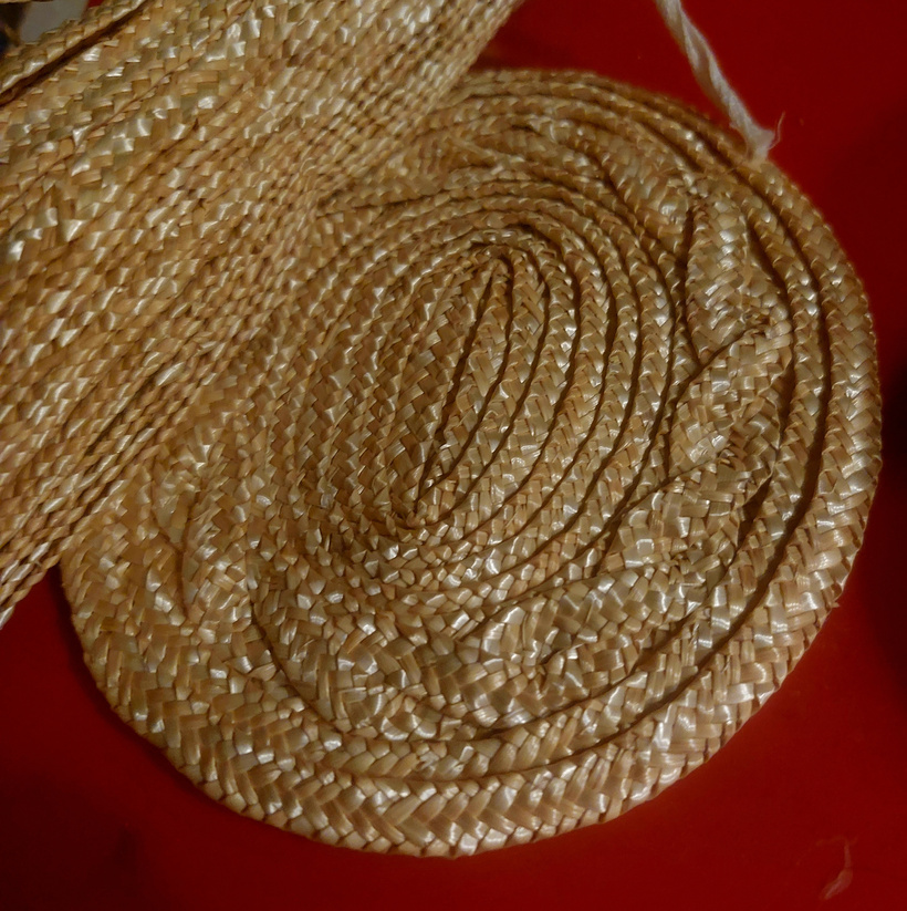 The start of a handsewn straw hat's crown with a hank of straw across the top left corner. This portion of the crowns tip shows some of the vining that adds visual texture to the hat.