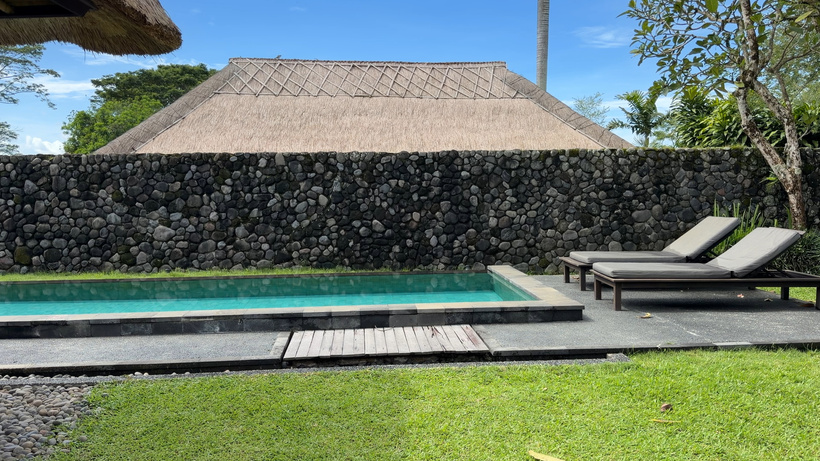 Two sun loungers beside a small pool in front of a river-stone wall and thatched roof of Pool Villa at Alila Ubud