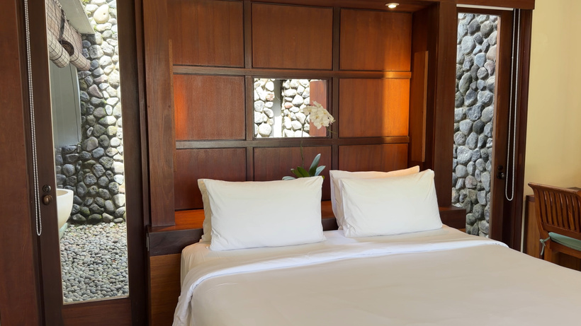 Double bed with white linen against a wood-panelled wall in the Deluxe room at Alila Ubud, with a stone wall visible through the window