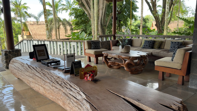 Reception seating area at Alila Ubud with cushioned sofas, carved wooden coffee tables and a rustic timber counter