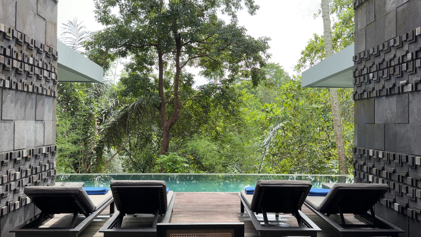 View between two dark stone pillars over sun loungers, a wooden deck and a private pool toward the jungle of 2 Bedroom Pool Villa at Alila Ubud