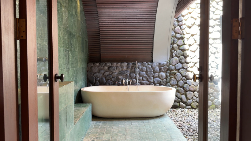 Freestanding white bathtub in the open-air bathroom of the Deluxe room at Alila Ubud, beside a river-stone wall and timber doors