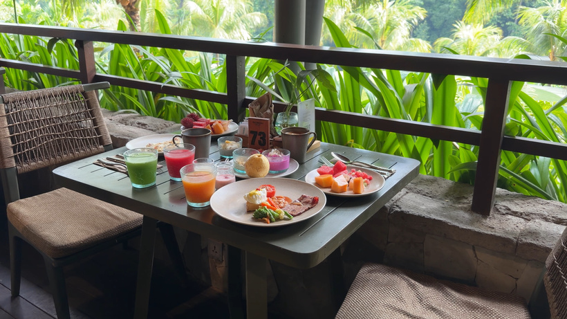 Outdoor breakfast table with plates of eggs and fruit and green, red and orange juices, by a railing with jungle views.