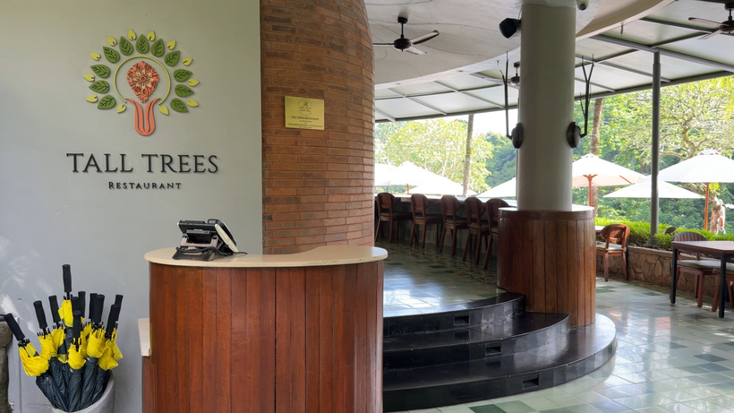 Tall Trees restaurant entrance with a wooden host stand, logo on the wall and an open-air dining area with parasols beyond.