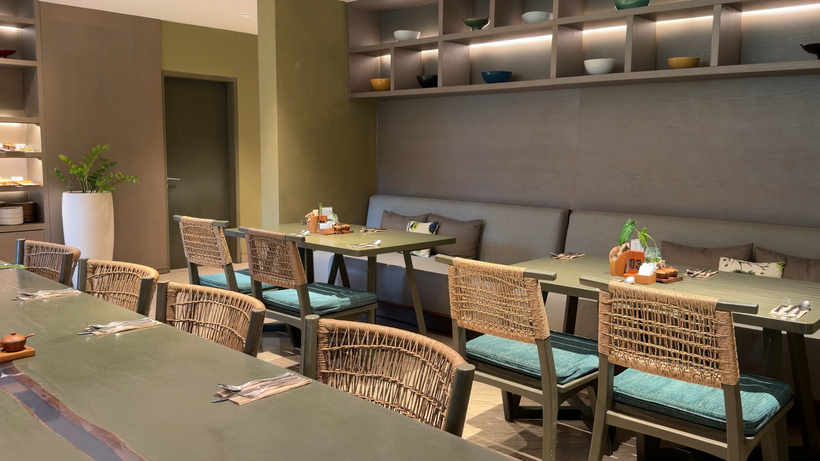 Indoor dining room with long tables, rattan chairs with teal cushions and a grey banquette under warm shelf lighting.