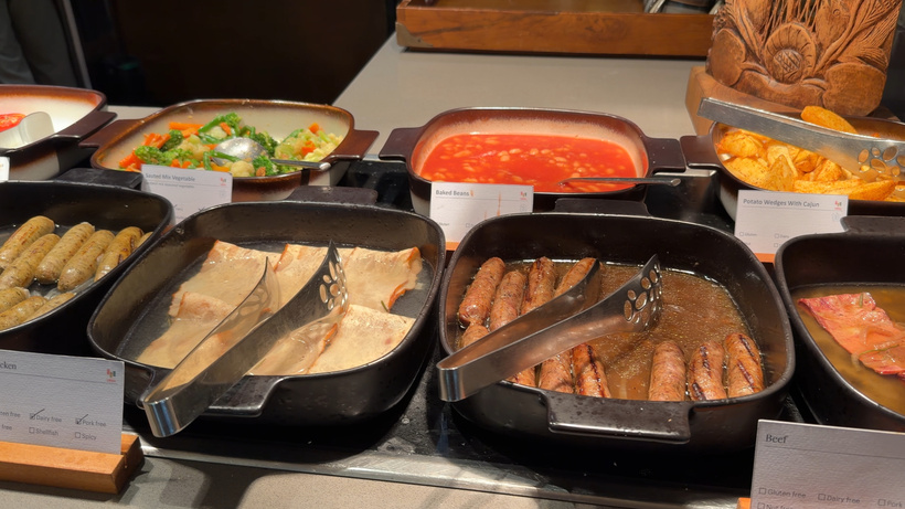 Hot buffet trays with fish fillets, sausages with tongs, a tomato dish and vegetables, each with a label card.
