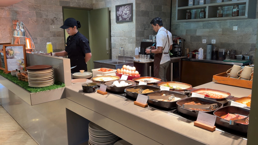 Two chefs at a live cooking station behind a buffet counter of hot dishes, eggs and stacked plates.