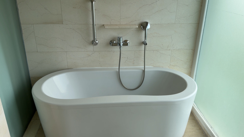 Freestanding white oval bathtub with a wall-mounted tap and handheld shower against cream stone tiles.