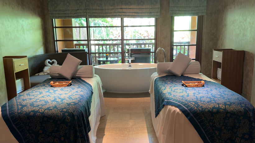 Spa treatment room with two massage beds in blue patterned covers and a freestanding tub by large windows.