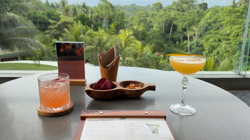 Table with an orange cocktail in a coupe glass, a pink drink, a bowl of snacks and a menu, with jungle behind.
