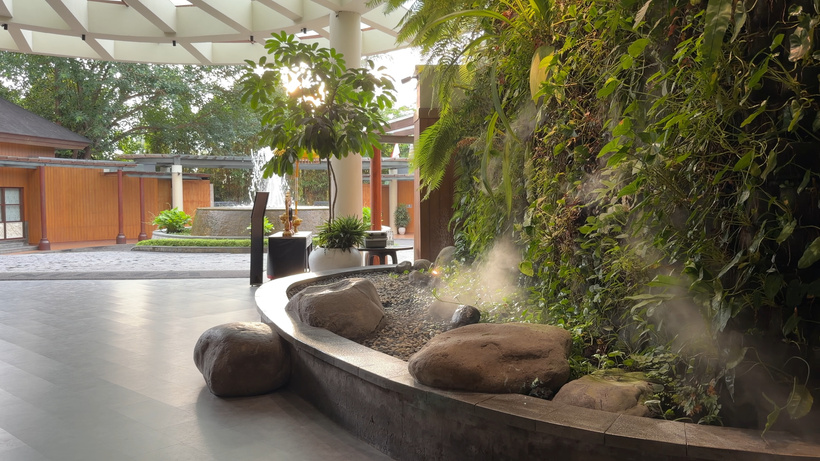 Open-air lobby entryway with a vertical moss and plant wall, decorative rock feature with mist, and sunlight filtering through the open entrance.   