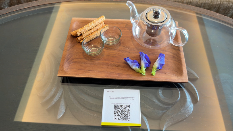 Walnut tray on a glass table with a glass teapot, two small cups, butterfly pea flowers, biscuit rolls and a QR code card.