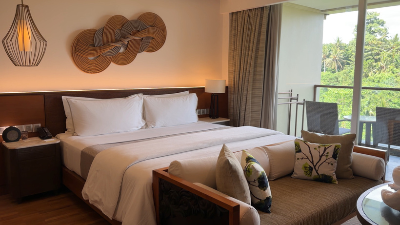 King bed with white linen and a cushioned sofa at its foot, with balcony doors and trees beyond.