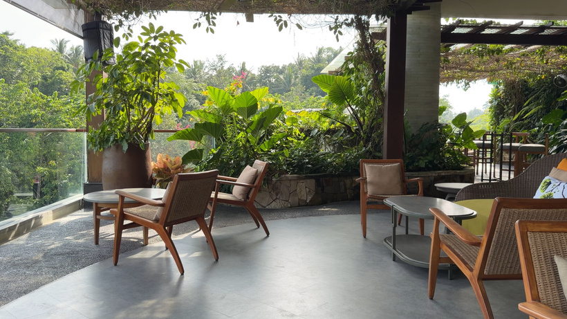 Open-air lounge with wooden armchairs, low tables and potted plants overlooking trees and the valley.