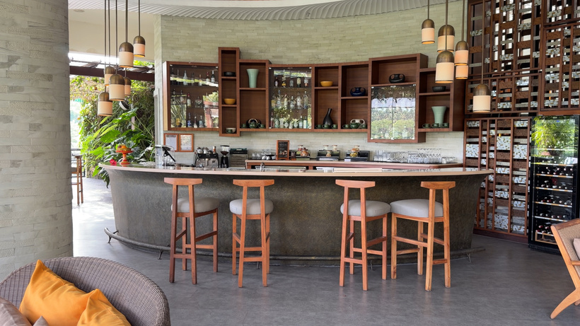 Curved bar with wooden stools under pendant lights, with shelves of glasses and a wine wall behind.