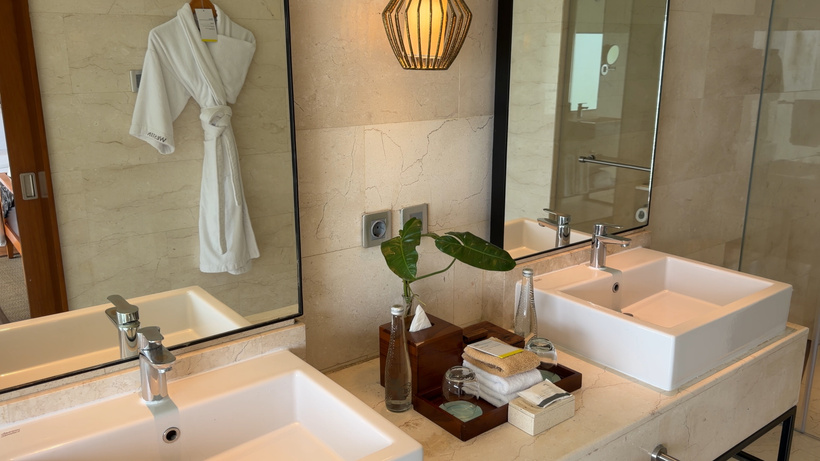 Double vanity with two white rectangular basins, mirrors, a small plant and a tray of toiletries, with a robe hanging behind.