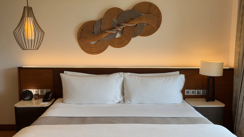 King bed with white pillows beneath a woven rope-style wall sculpture, with a pendant lamp and bedside lamps.