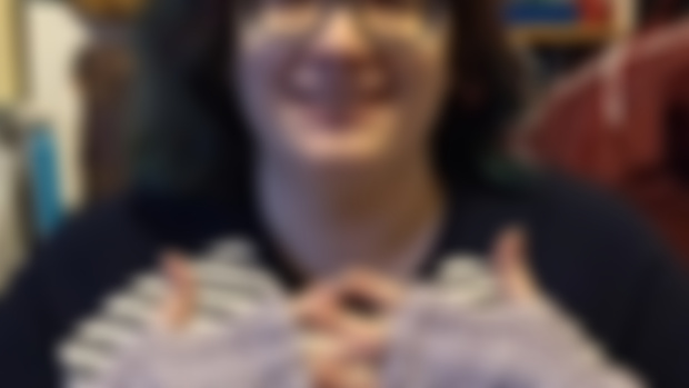 A white genderqueer woman (me) smiling at the camera with my hands in front of my chest. I'm wearing my Swift & Simple Lace Mitts, made in lilac-coloured yarn.